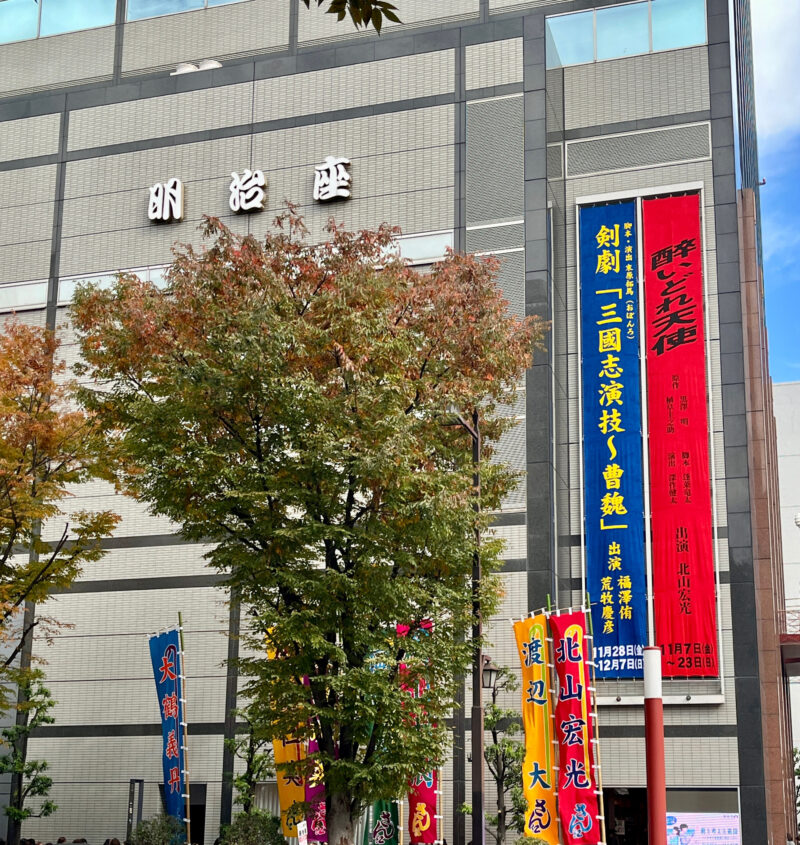 明治座の外観。建物の壁面に公演の縦看板が掲出され、前にはカラフルなのぼり旗と木々が見える。