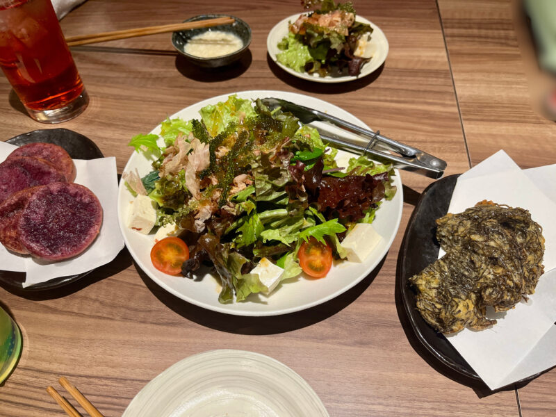 沖縄料理の前菜いろいろ。サラダ、もずくの天ぷら、紅いもチップスなどがテーブルに並んでいる。