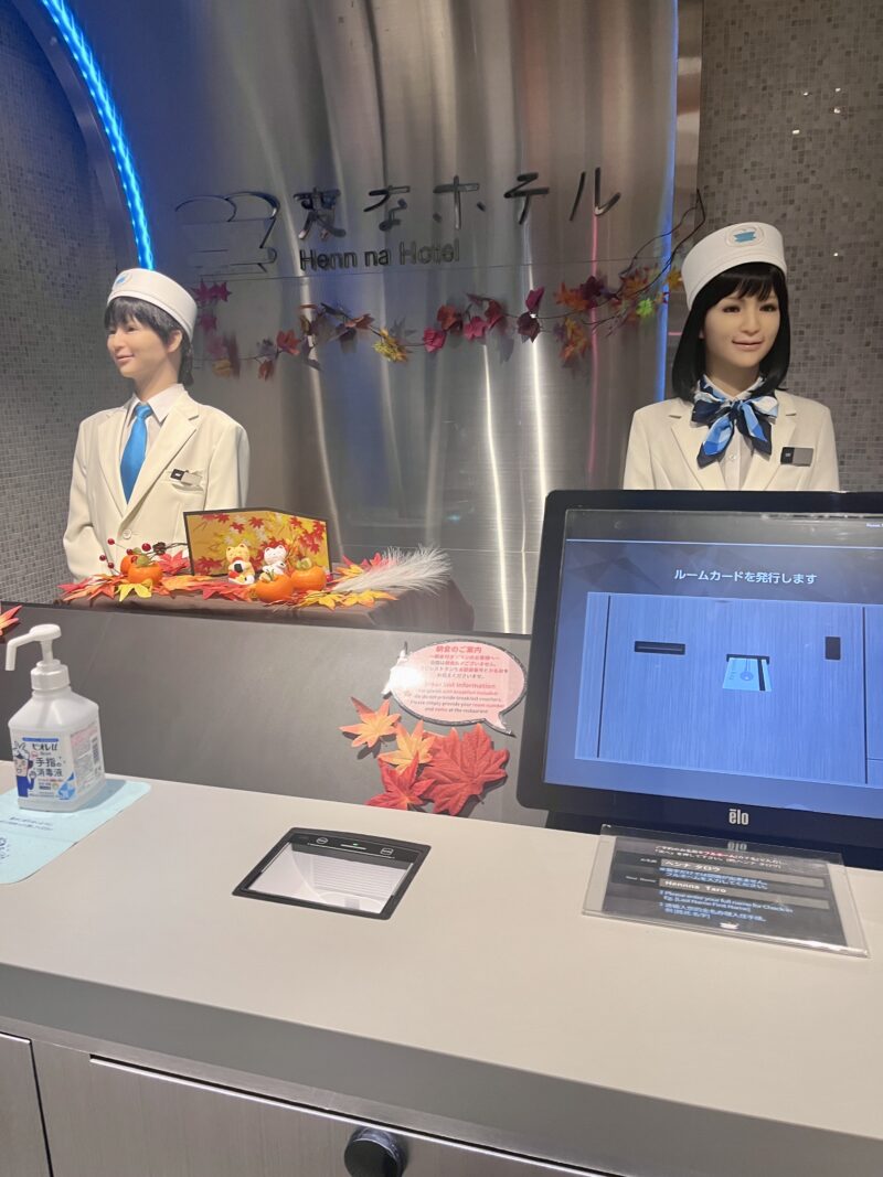 変なホテル東京浜松町のフロント写真。2体のヒューマノイドが並び、手前にはチェックイン用タッチパネルが設置されている。秋の装飾が施されたカウンター。
